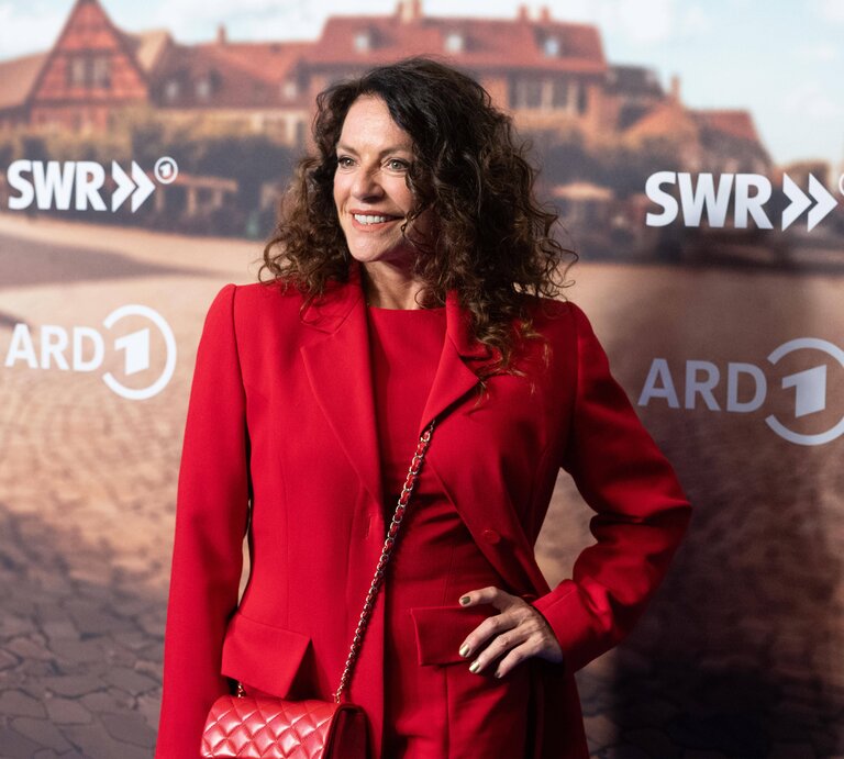 Christine Neubauer mit dunklen lockigen Haaren trägt ein elegantes rotes Outfit, dazu goldenen Nagellack und eine passende rote Tasche vor einem städtischen Hintergrund. | © IMAGO / Sven Simon