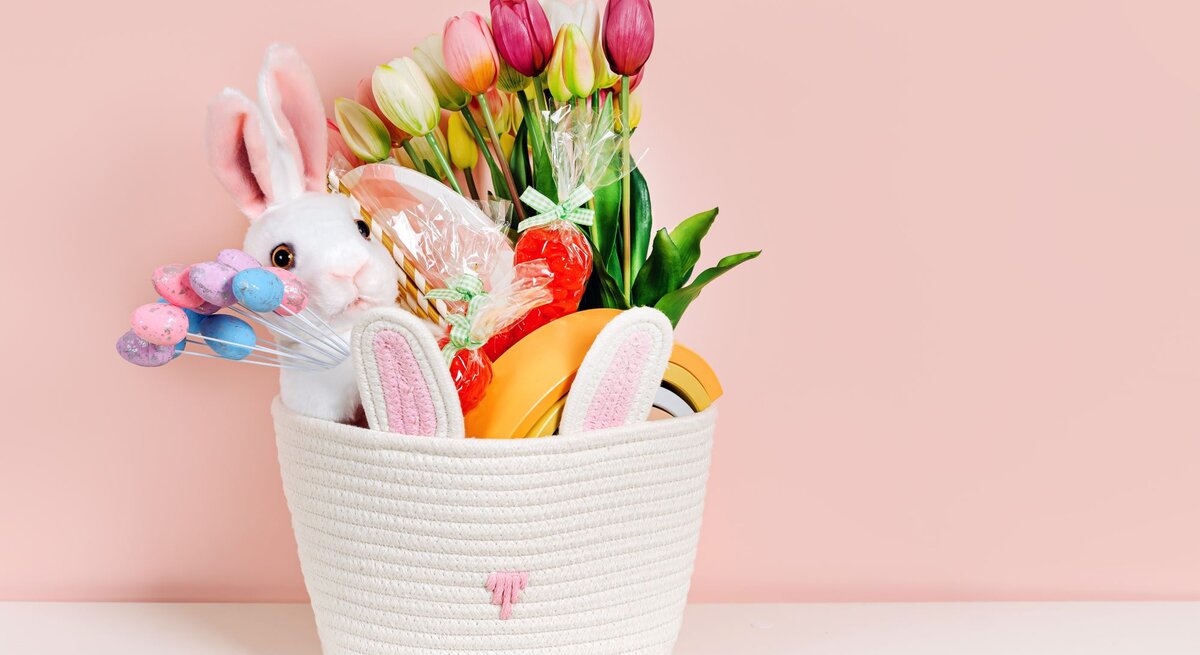Weißer Osterkorb mit Plüschhase, Tulpen, bunten Eiern und Süßigkeiten vor rosa Hintergrund auf weißer Fläche.