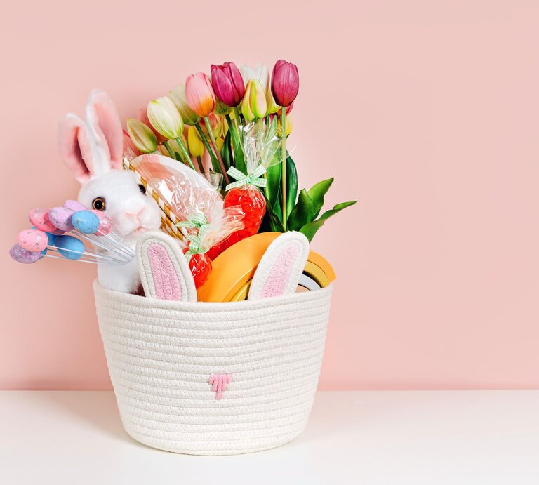 Weißer Osterkorb mit Plüschhase, Tulpen, bunten Eiern und Süßigkeiten vor rosa Hintergrund auf weißer Fläche. | © Shutterstock/ Igisheva Maria