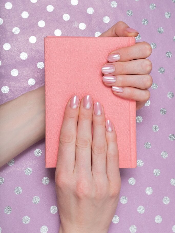 Eine Hand mit elegantem, nudefarbenem Nagellack und pinkem French-Tip trägt einen glänzenden goldenen Ring und liegt auf einem grauen Blazer. | © FreePik/ somemeans