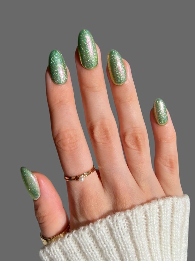 Eine Hand mit glänzenden, grünen Glitzer-Nägeln trägt zwei goldene Ringe, darunter ein Ring mit Perle, und steckt in einem weißen Strickpulloverärmel. | © Adobe Stock/ Max
