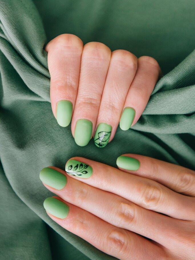 Zwei Hände mit mattem, grünen Nagellack und einem Blattmuster auf dem Ringfinger liegen nebeneinander auf einem grünen Stoff. | © Adobe Stock/ Alex Photo