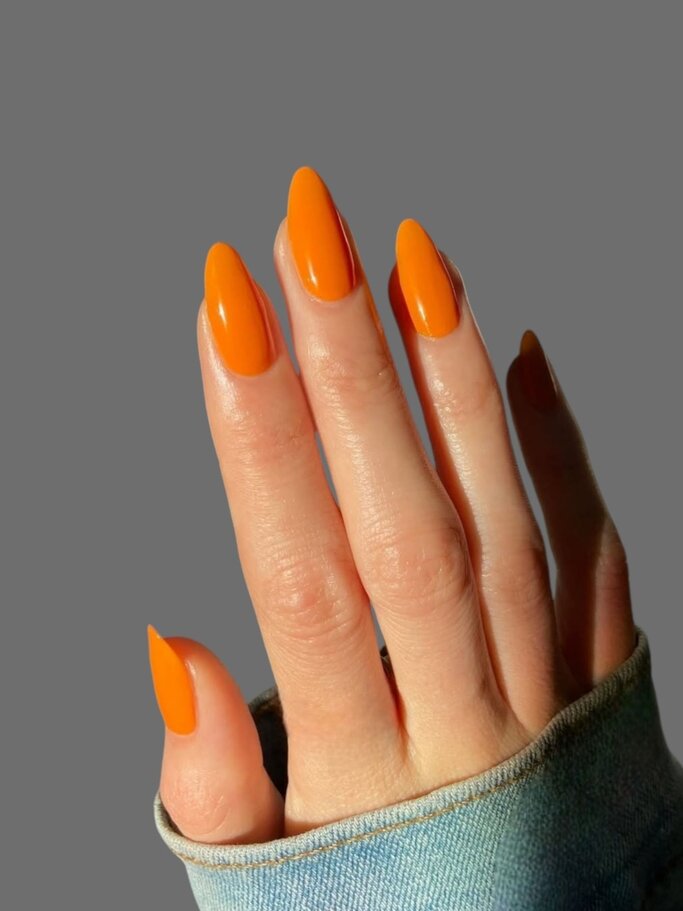 Nahaufnahme einer Hand mit langen, mandelförmigen Nägeln in glänzendem Orange vor grauem Hintergrund. | © Adobe Stock/ Max