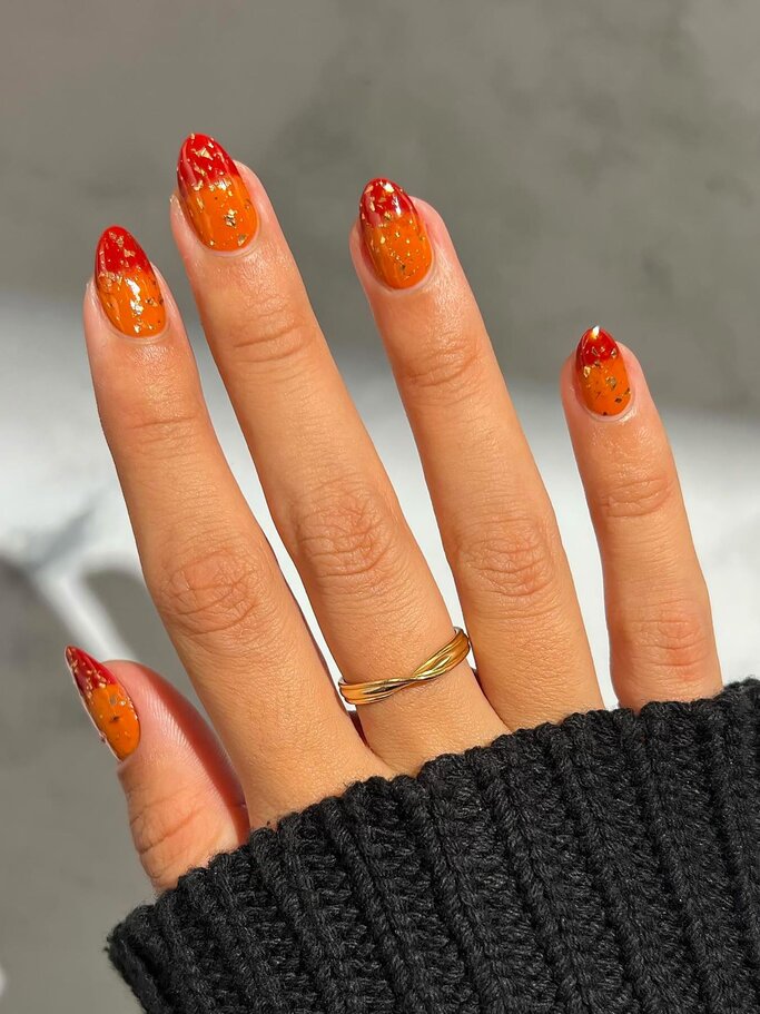 Eine Hand mit leuchtend orange-roten Nägeln und goldenen Verzierungen trägt einen goldenen Ring und steckt in einem schwarzen, gestrickten Ärmel. | © Shutterstock/ Art of Body