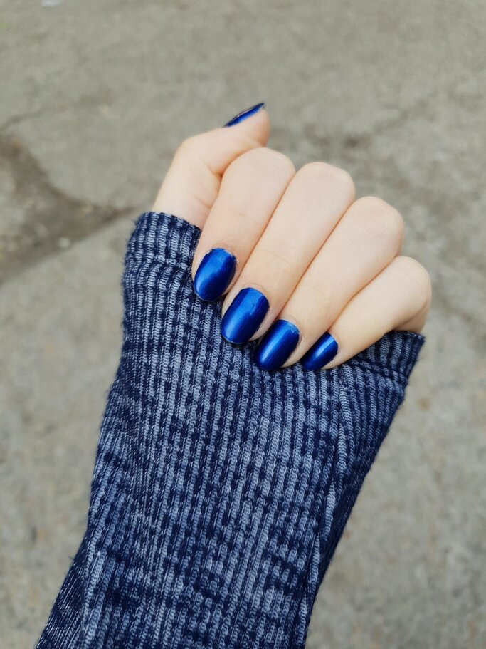 Eine Hand mit glänzend blauen Nägeln trägt einen grauen, gestrickten Pulloverärmel und ist vor einem unscharfen grauen Hintergrund abgebildet. | © FreePik/ EyeEm