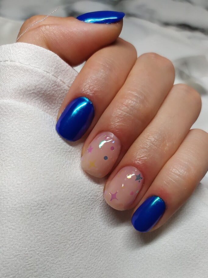 Eine Hand mit glänzendem blauen Nagellack und zwei funkelnden, mit bunten Sternen verzierten Nägeln, die in einem weißen Stoff eingefasst sind. | © Getty Images/ mikroman6