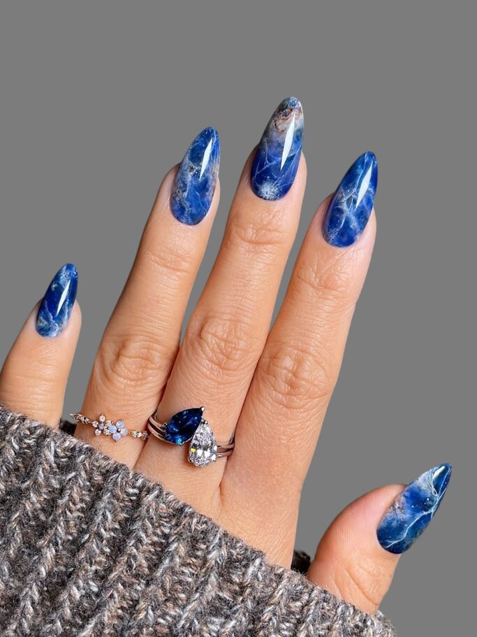 Eine Hand mit langen, spitzen blauen Nägeln im Marmor-Look trägt zwei elegante Ringe und steckt in einem grauen Strickpullover. | © Getty Images/ Marine Saad