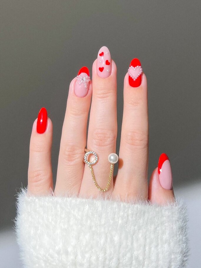Eine Hand mit rot-rosa Nageldesign, verziert mit Herzen und Perlen, trägt einen eleganten Ring mit einer Kette und einer Perle. | © Shutterstock/ Julian Photo Nail