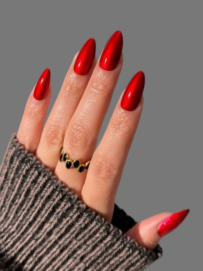 Eine Hand mit spitzen, glänzend roten Nägeln trägt einen goldenen Ring mit schwarzen Edelsteinen, umhüllt von einem grauen Strickpullover. | © Adobe Stock/ Max