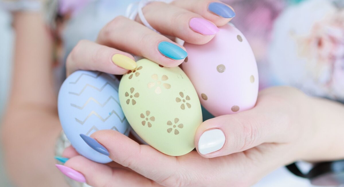 Hände einer jungen Frau mit pastellfarbenem Nageldesign halten drei bemalte Ostereier mit goldenen Mustern vor bunter Kleidung.