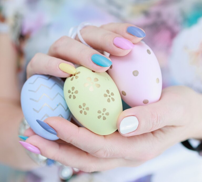 Hände einer jungen Frau mit pastellfarbenem Nageldesign halten drei bemalte Ostereier mit goldenen Mustern vor bunter Kleidung. | © Shutterstock/ TanitaKo