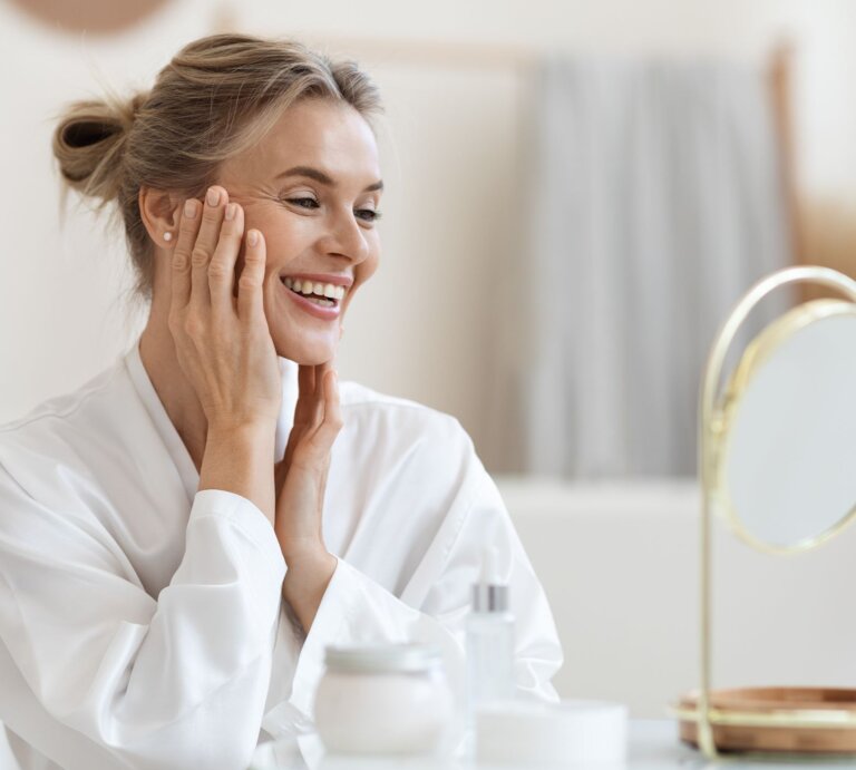Frau in weißem Bademantel berührt sanft ihr Gesicht, vor einem Tisch mit Creme und einem goldenen Kosmetikspiegel im hellen Raum. | © Adobe Stock/ Prostock-studio