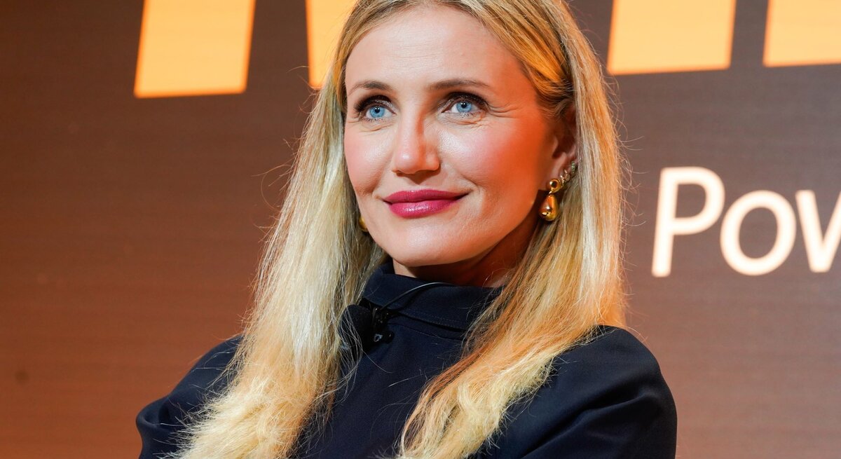 Cameron Diaz mit langen, offenen Haaren und goldenen Ohrringen lächelt dezent vor einem orange-schwarzen Hintergrund.