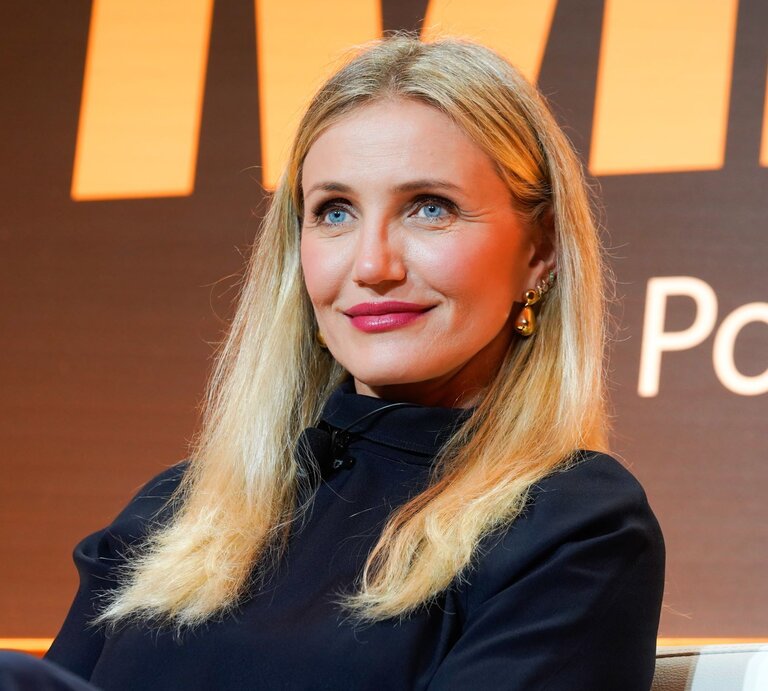 Cameron Diaz mit langen, offenen Haaren und goldenen Ohrringen lächelt dezent vor einem orange-schwarzen Hintergrund. | © Presley Ann/Getty Images for Fortune's Most Powerful Women Summit