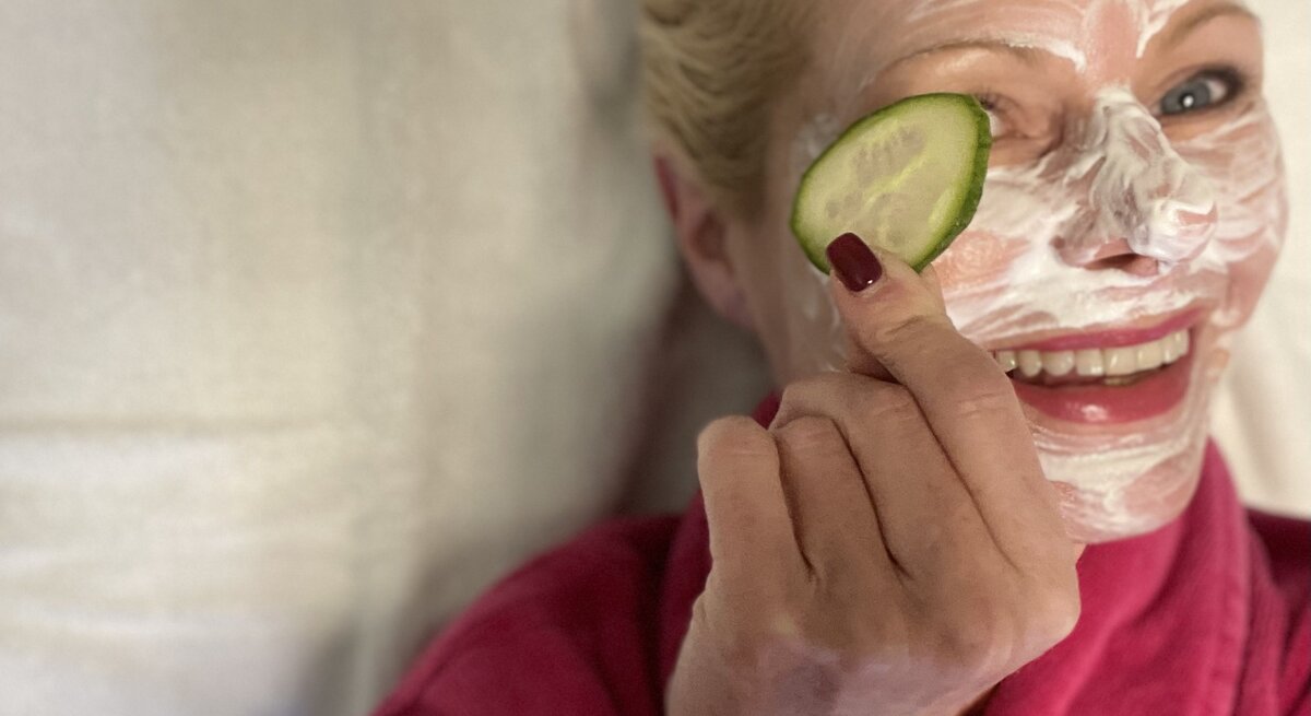 Eine lächelnde blonde Frau mittleren Alters in einem pinken Bademantel trägt eine Gesichtsmaske und hält eine Gurkenscheibe vor ihr Auge.