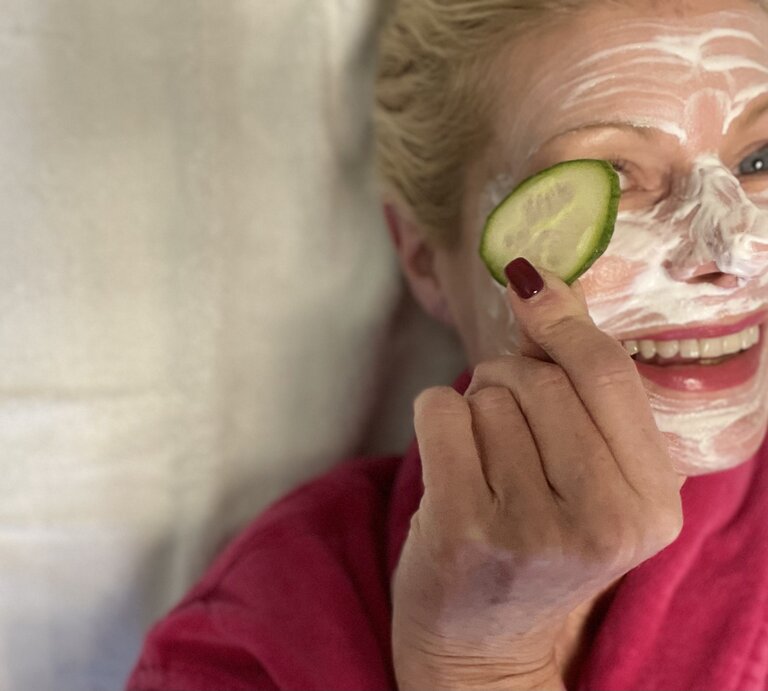 Eine lächelnde blonde Frau mittleren Alters in einem pinken Bademantel trägt eine Gesichtsmaske und hält eine Gurkenscheibe vor ihr Auge. | © Renate Zott
