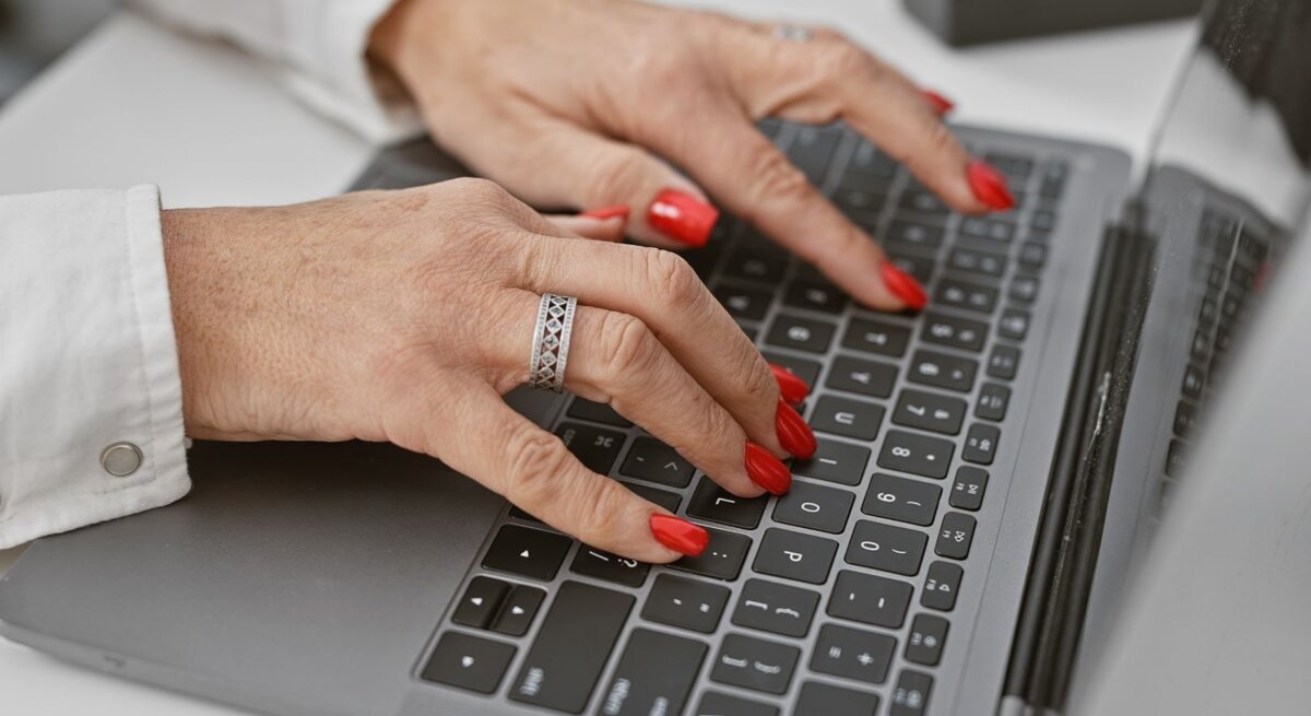 Gepflegte Frauenhände mit rotem Nagellack und silbernem Ring tippen auf einer Laptop-Tastatur in modernem Büroambiente.