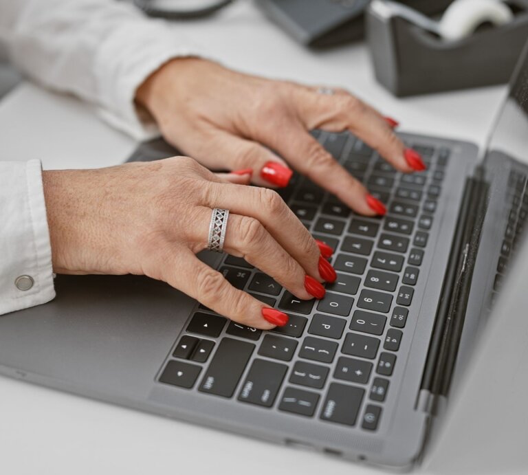 Gepflegte Frauenhände mit rotem Nagellack und silbernem Ring tippen auf einer Laptop-Tastatur in modernem Büroambiente. | © Shutterstock / Krakenimages.com