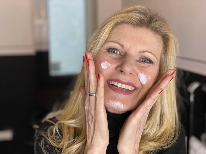 Lachende blonde Frau mittleren Alters trägt Gesichtscreme auf den Wangen, hat rote Fingernägel und stützt ihr Gesicht mit den Händen. | © Renate Zott