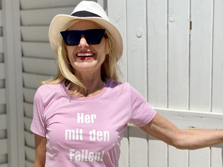 Lachende Frau mit Sonnenbrille und Hut, die ein rosa T-Shirt mit dem Aufdruck „Her mit den Falten!“ trägt.  | © Renate Zott