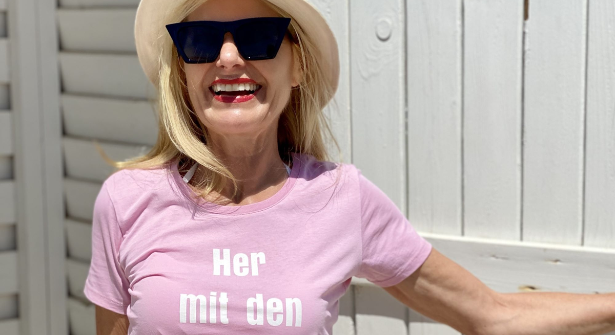 Lachende Frau mit Sonnenbrille und Hut, die ein rosa T-Shirt mit dem Aufdruck „Her mit den Falten!“ trägt. 