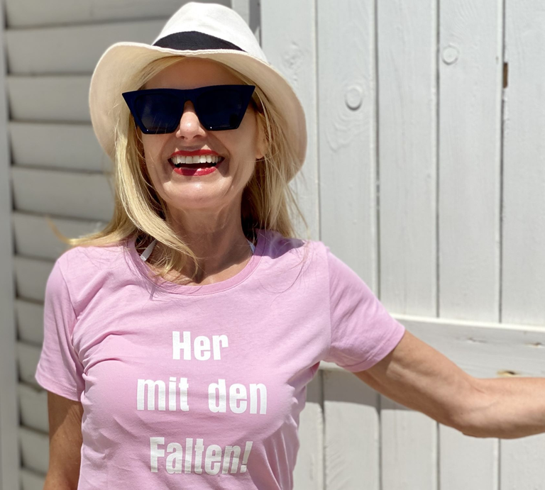 Lachende Frau mit Sonnenbrille und Hut, die ein rosa T-Shirt mit dem Aufdruck „Her mit den Falten!“ trägt.  | © Renate Zott