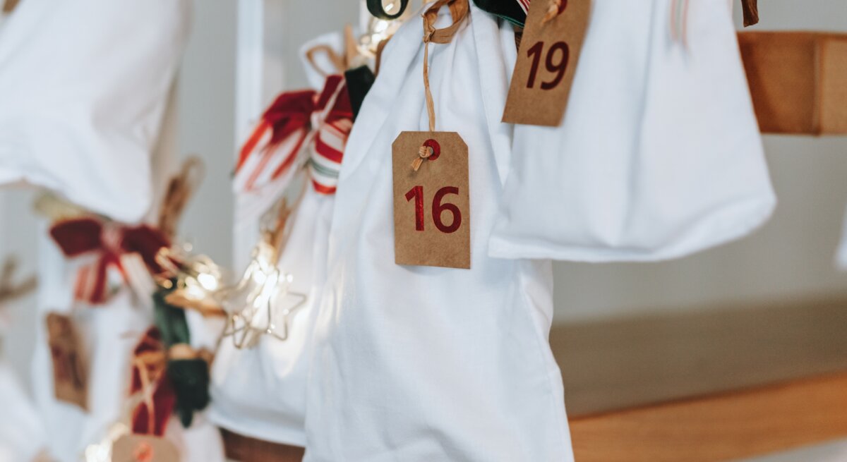 Weiße Stoffbeutel mit roten Zahlen und Schleifen hängen als dekorativer Adventskalender an einer Holztreppe, sanft beleuchtete Sterne.