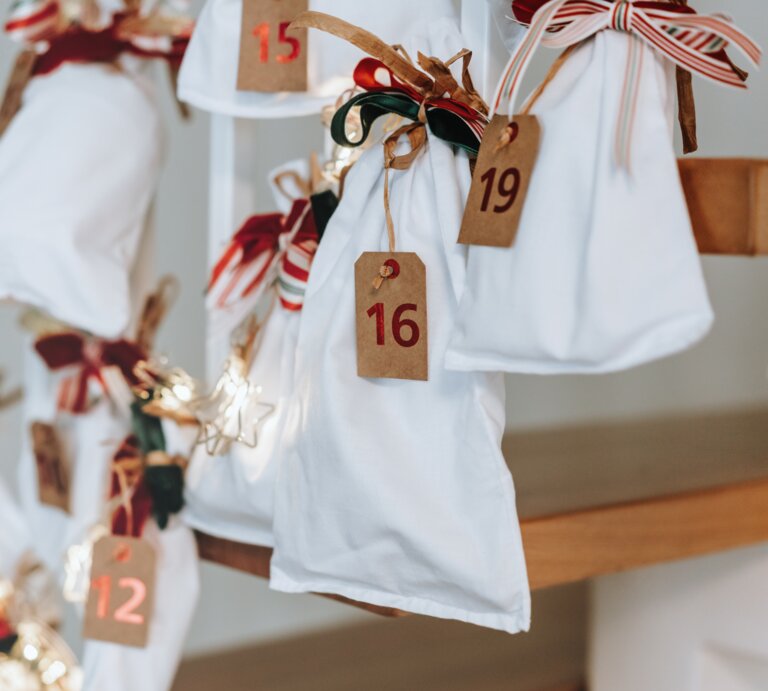 Weiße Stoffbeutel mit roten Zahlen und Schleifen hängen als dekorativer Adventskalender an einer Holztreppe, sanft beleuchtete Sterne. | © Adobe Stock/ RasaBasa