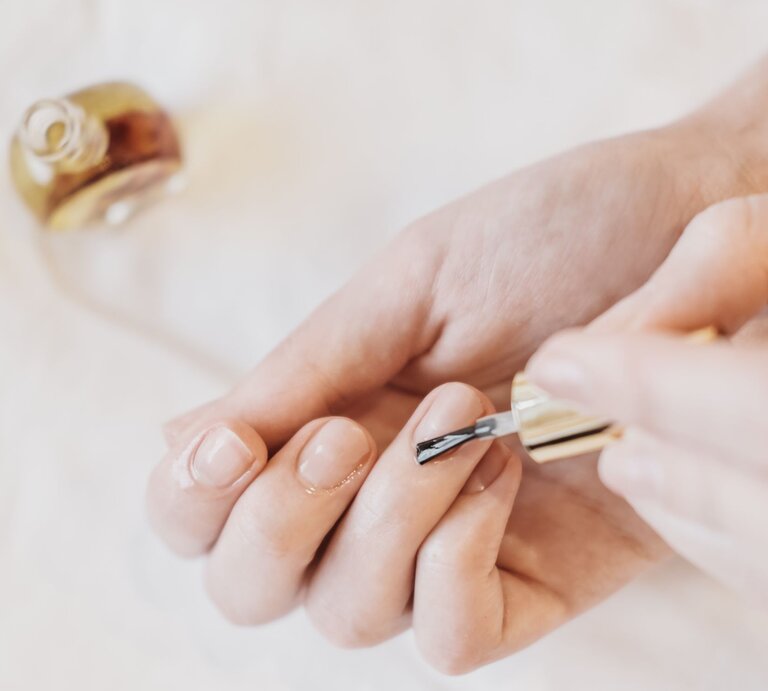 Eine weibliche Hand lackiert behutsam einen Fingernagel mit einem klaren Nagellack, im Hintergrund steht eine geöffnete Flasche. | © Adobe Stock/ Yulia Panova