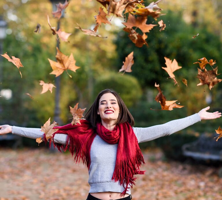 Eine junge Frau mit schulterlangem braunem Haar und rotem Lippenstift wirft lachend Herbstblätter in die Luft, umgeben von buntem Laub in einem Park. | © Adobe Stock/ tunedin