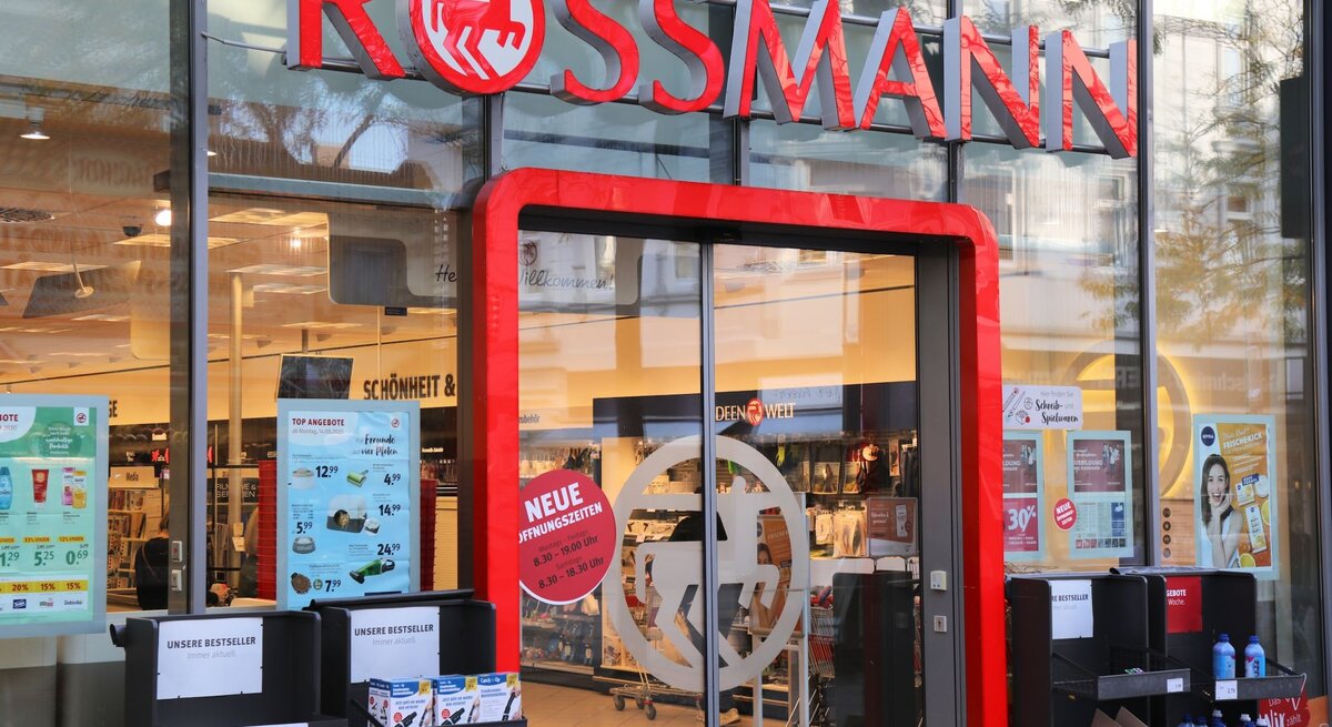 Frontansicht eines Drogeriemarktes von Rossmann mit Schaufensterplakaten, Einkaufswagen und Regalen mit Produkten vor der Tür.