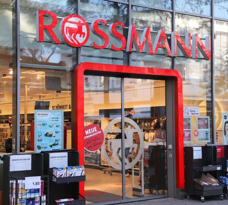 Frontansicht eines Drogeriemarktes von Rossmann mit Schaufensterplakaten, Einkaufswagen und Regalen mit Produkten vor der Tür. | © Adobe Stock/ Tupungato