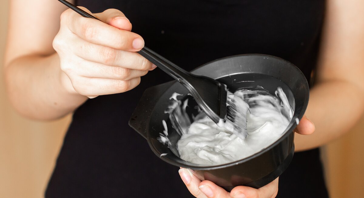 Hände einer Frau halten eine schwarze Schale mit cremiger Haarfarbe, während sie die Farbe mit einem schwarzen Pinsel rührt.