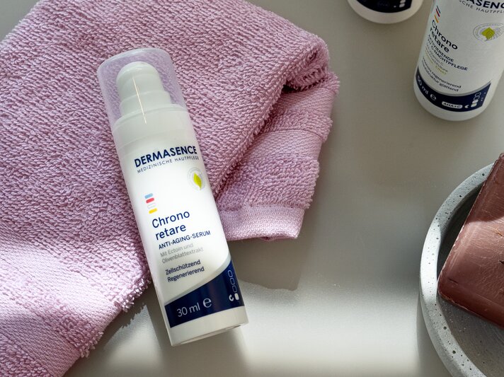 Das Dermasence Chrono Retare Anti-Aging-Serum liegt auf einem rosa Handtuch, daneben ein Seifenstück auf einem Teller. | © Dermasence