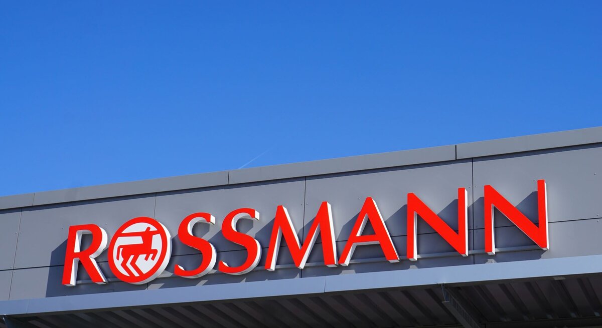 Rotes Rossmann-Logo auf grauer Fassade eines Geschäftes vor klarem, blauem Himmel.