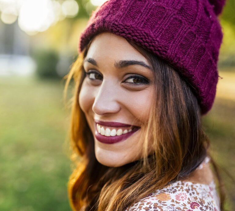 Junge Frau mit lebendigem Lächeln und bordeauxfarbener Strickmütze in herbstlicher Parklandschaft im goldenen Licht. | © Getty Images/ Westend61
