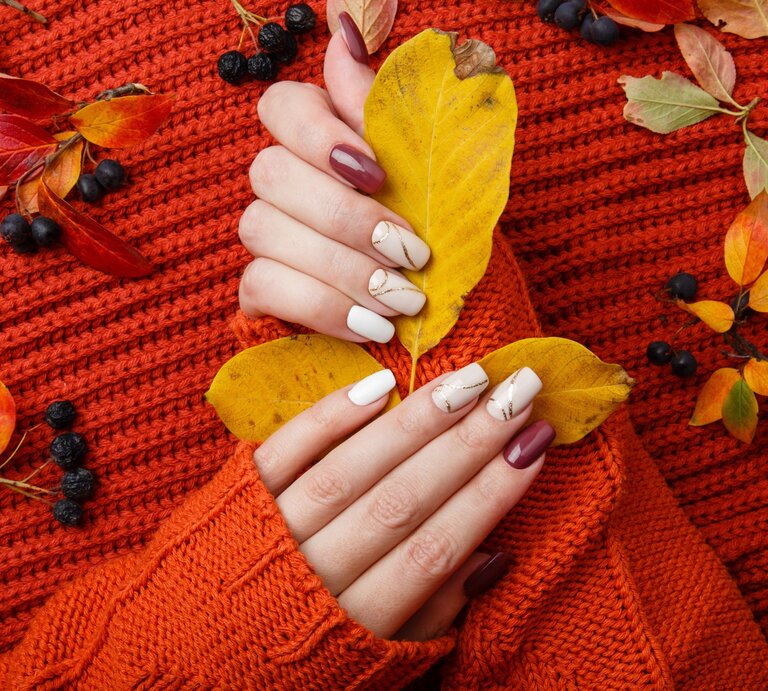 Hände mit herbstlich bemalten Nägeln halten gelbe Blätter auf einem orangefarbenen Strickpullover, umgeben von buntem Herbstlaub | © Adobe Stock/ Olga Mishyna