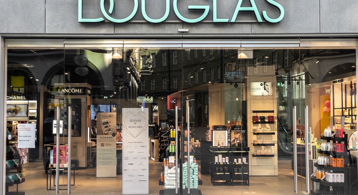 Eingang des Douglas-Geschäfts mit leuchtendem Logo, umgeben von Kosmetik-Displays und Werbung.
