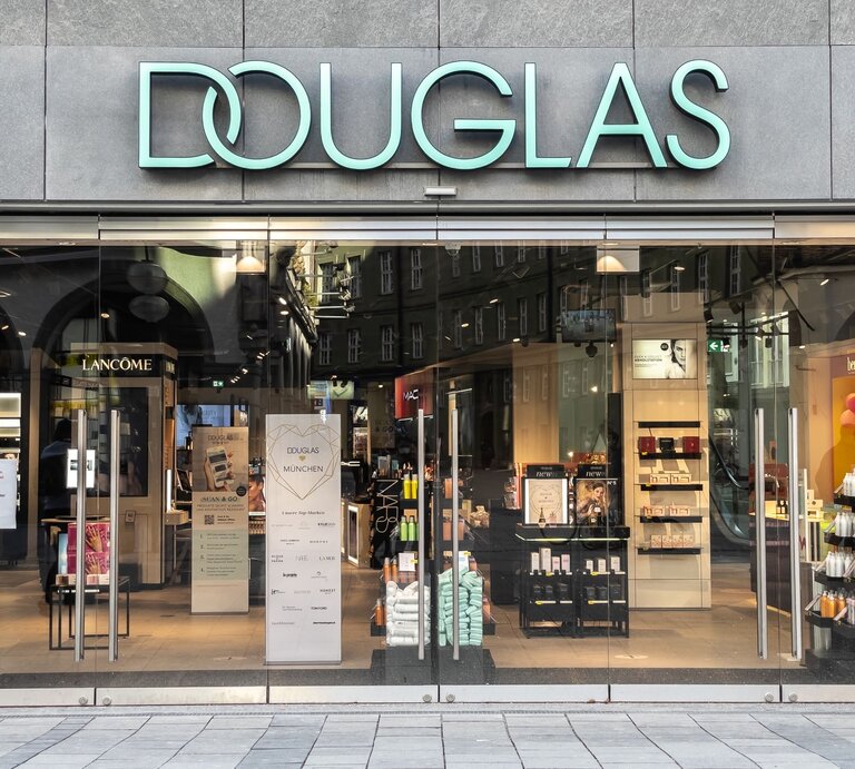 Eingang des Douglas-Geschäfts mit leuchtendem Logo, umgeben von Kosmetik-Displays und Werbung. | © Adobe Stock/ Dennis