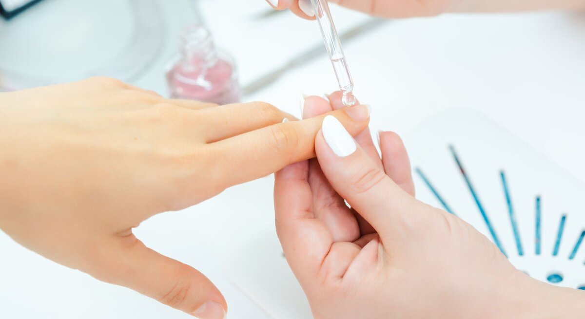 Nahaufnahme einer klassischen Maniküre: Nagelöl wird präzise mit einer Tropfpipette auf den Nagel einer Hand aufgetragen.