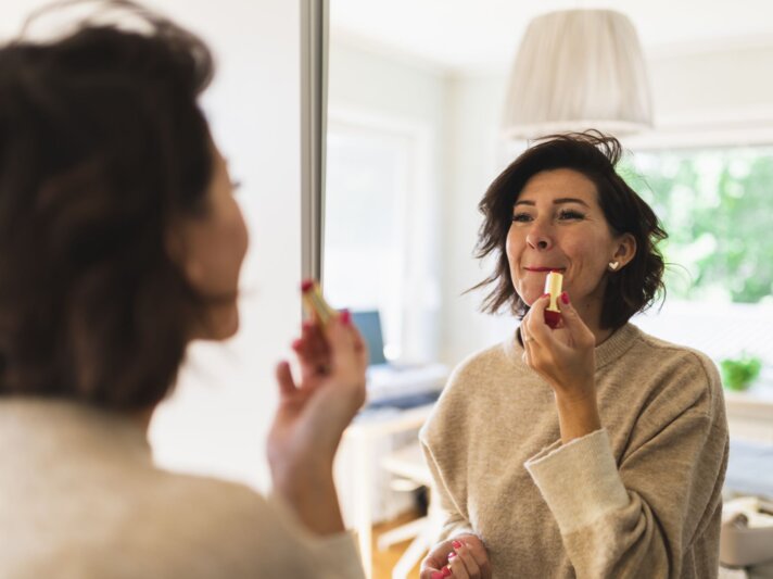 Frau im beigen Pullover trägt Lippenstift vor einem Spiegel auf, im Hintergrund ein gemütliches, gut beleuchtetes Wohnzimmer. | © Getty Images/ Westend61