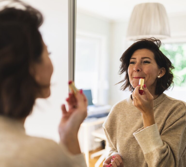Frau im beigen Pullover trägt Lippenstift vor einem Spiegel auf, im Hintergrund ein gemütliches, gut beleuchtetes Wohnzimmer. | © Getty Images/ Westend61