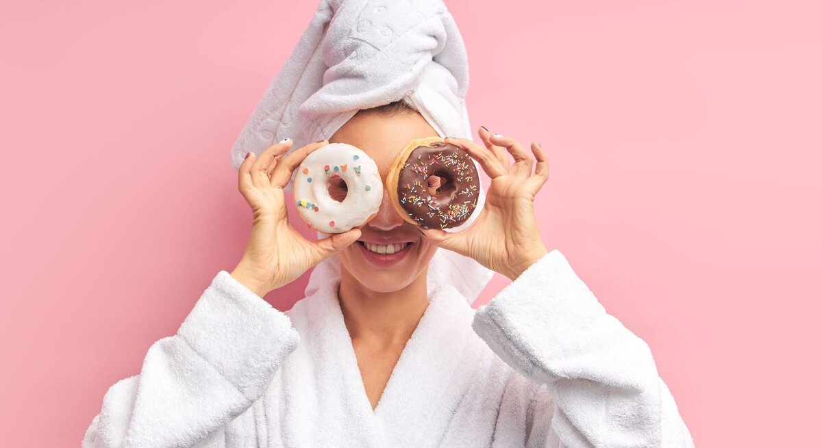Frau in Bademantel und Handtuch lacht und hält Donuts mit Glasur und Streuseln vor ihren Augen vor rosa Hintergrund.