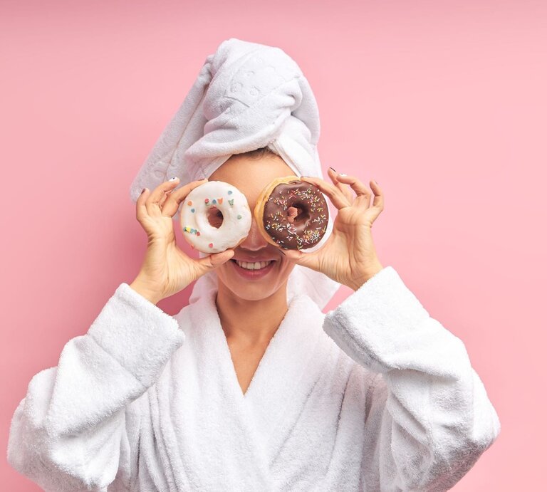Frau in Bademantel und Handtuch lacht und hält Donuts mit Glasur und Streuseln vor ihren Augen vor rosa Hintergrund. | © Adobe Stock/ alfa27