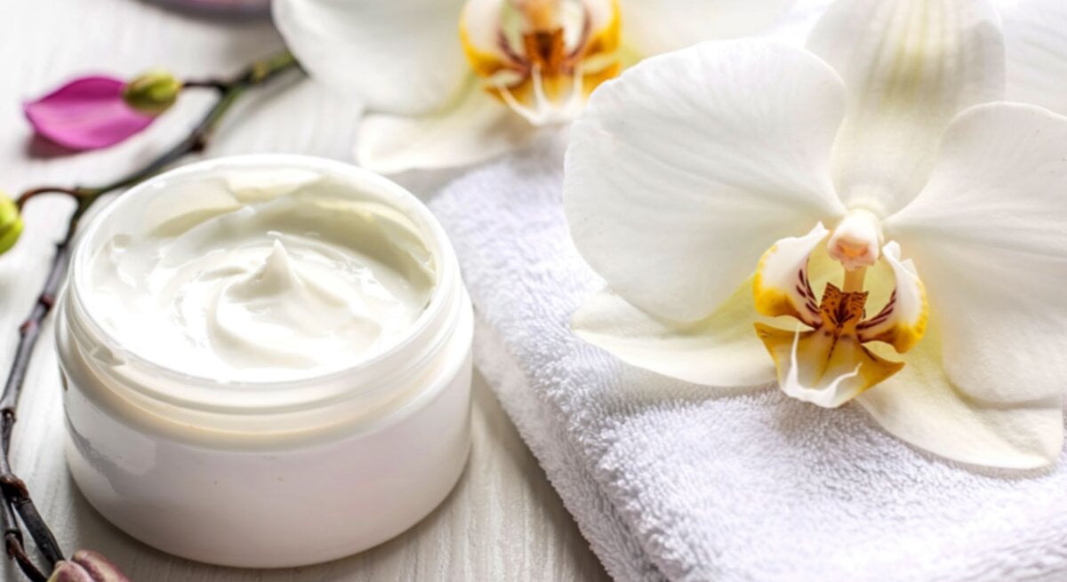 Eine luxuriöse Anti-Aging-Creme in Glasdose liegt auf einem weißen Handtuch, umgeben von Orchideen und Blütenblättern.