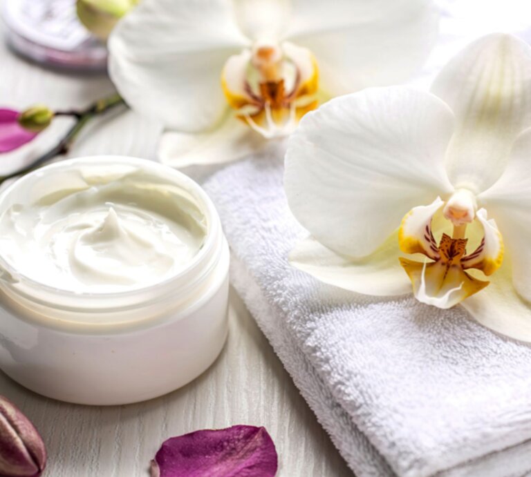 Eine luxuriöse Anti-Aging-Creme in Glasdose liegt auf einem weißen Handtuch, umgeben von Orchideen und Blütenblättern. | © Shutterstock/ RogozVasile