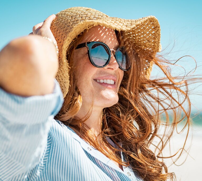 Rothaarige Frau in gestreifter Bluse genießt den Tag am Meer, eingecremt mit Sonnencreme und lächelt in die Sonne, während ihr Haar lebhaft flattert. | © Shutterstock/ Ground Picture