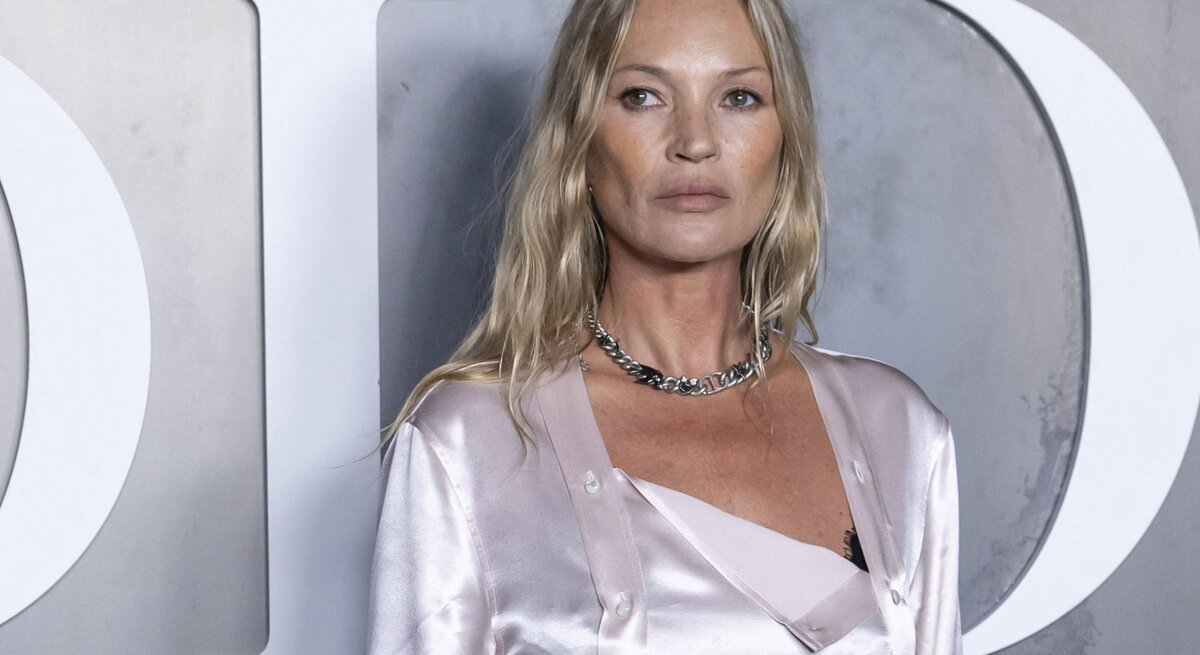 Kate Moss mit welligem Haar trägt ein seidiges, hellrosafarbenes Outfit und eine silberne Kette vor grauem Hintergrund.