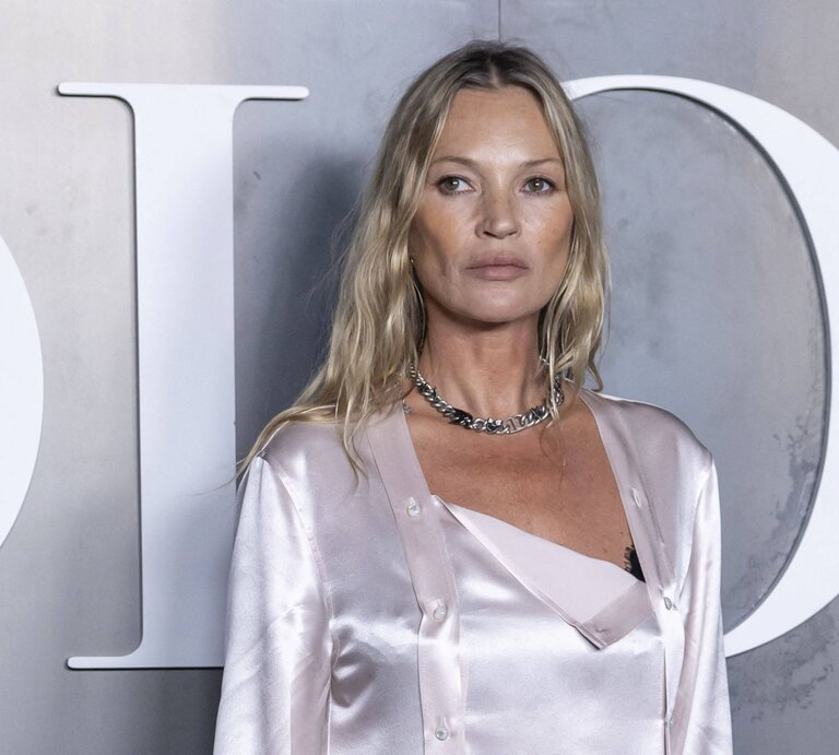 Kate Moss mit welligem Haar trägt ein seidiges, hellrosafarbenes Outfit und eine silberne Kette vor grauem Hintergrund. | © IMAGO / Bestimage