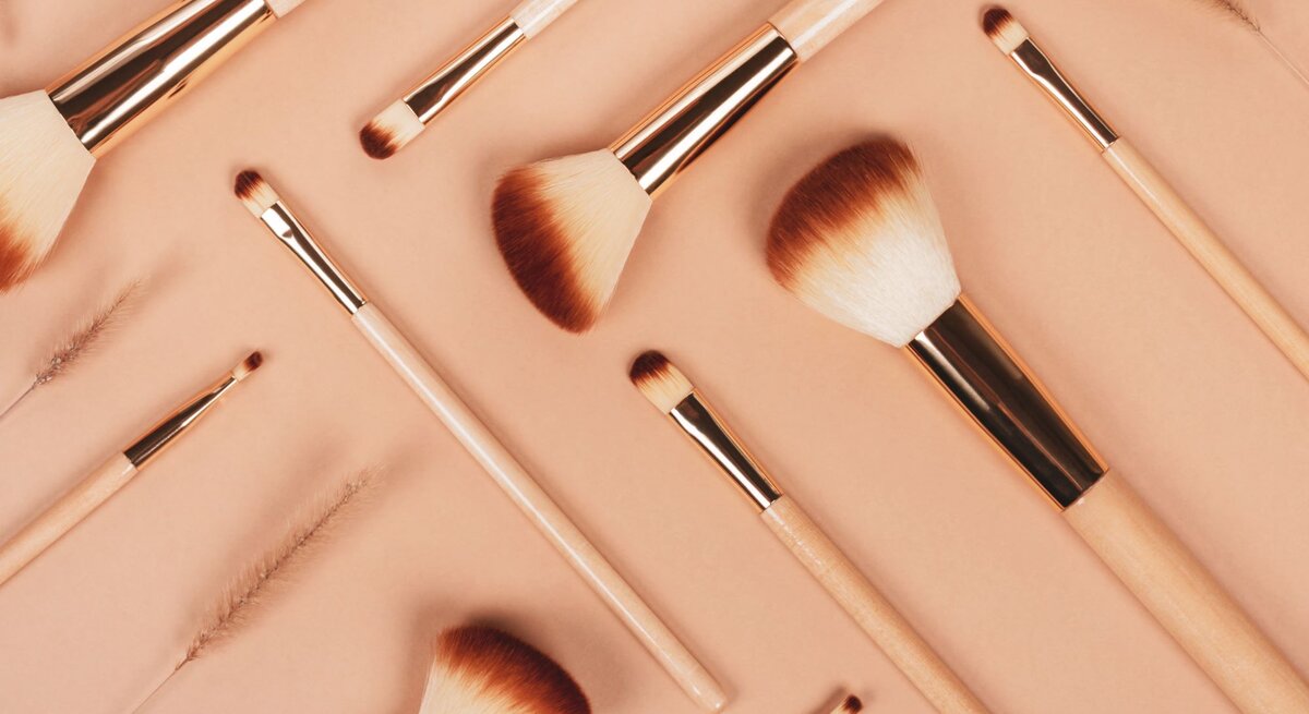 Verschiedene Make-up-Pinsel mit hellen Holzgriffen und braunen Borsten liegen ordentlich auf einem beigen Untergrund.